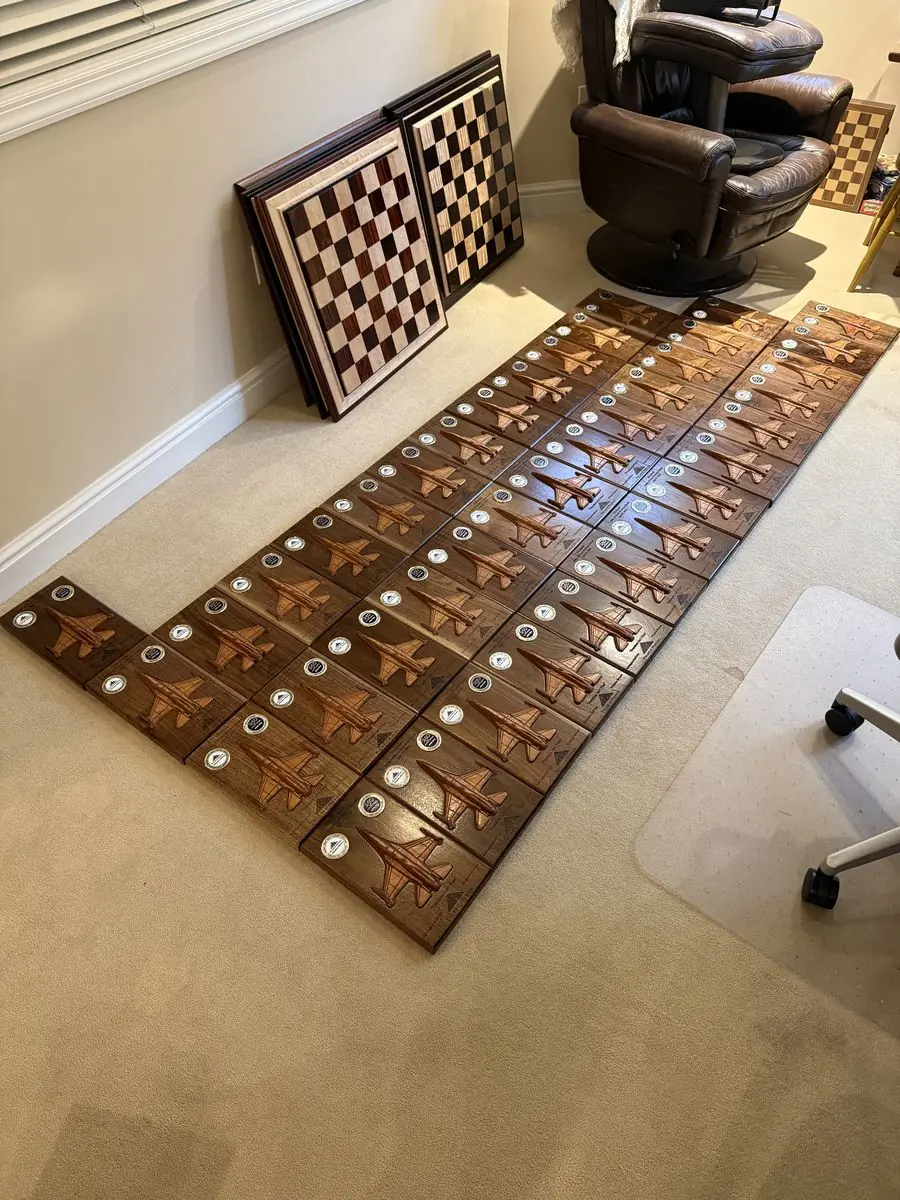 Rows of dark wood plaques laid out on a carpet, each with a carved jet and a challenge coin, with two chessboards leaning on the wall behind