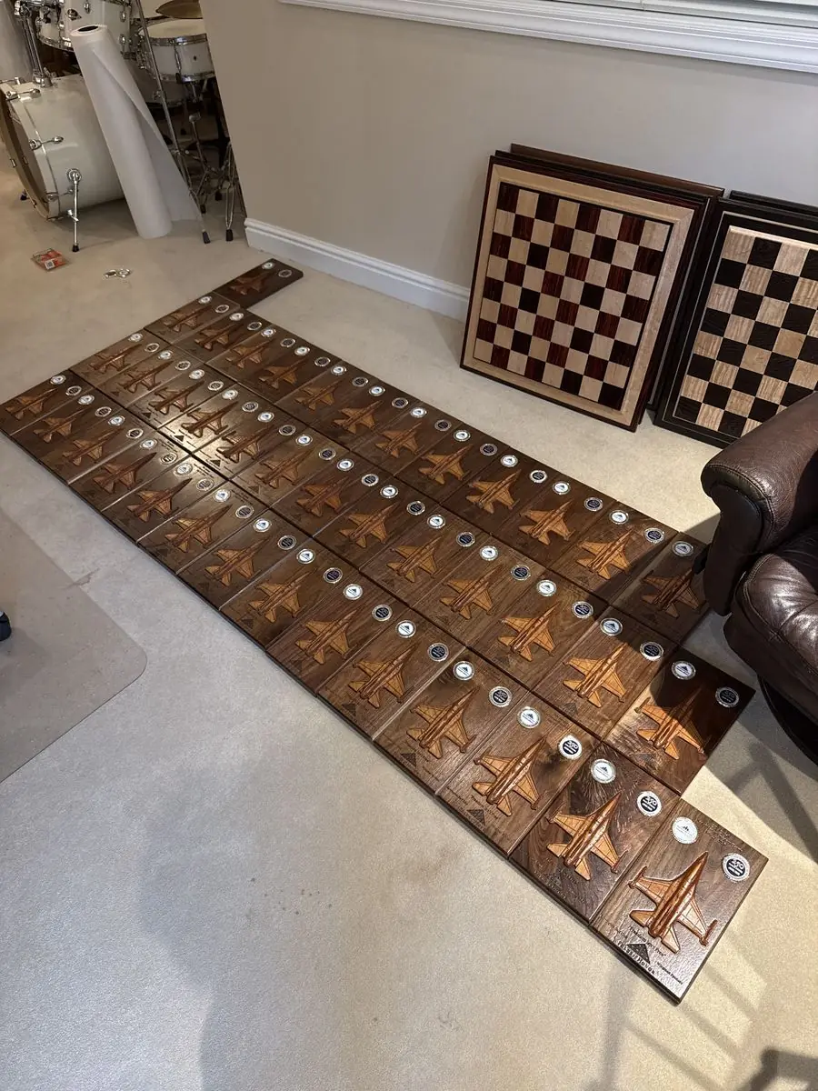 Rows of dark wood plaques with carved jets and challenge coins laid out on a carpet, with two chessboards leaning on the wall