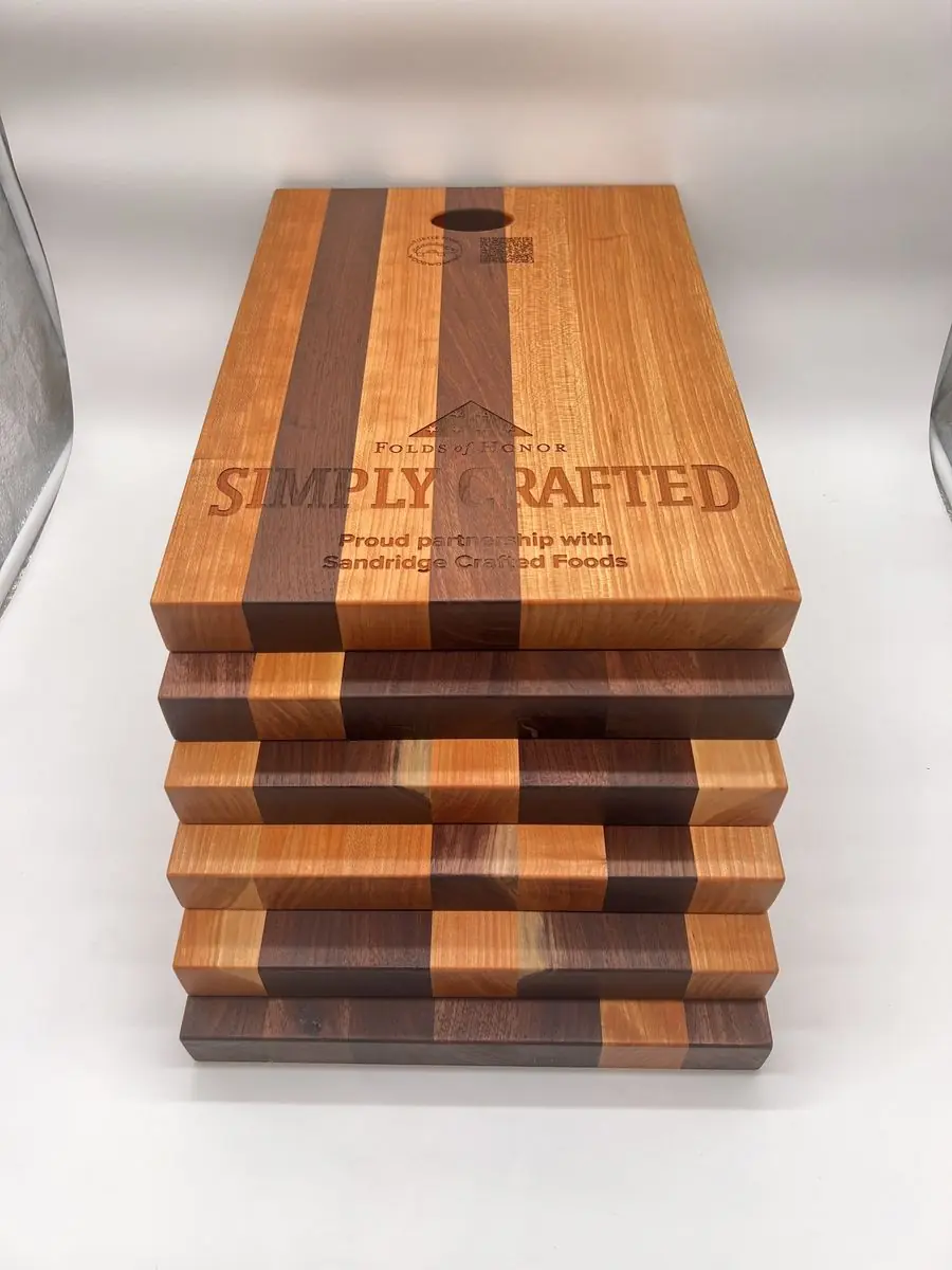 Stack of six cutting boards with alternating light and dark wood stripes and a finger hole; the top board is engraved with logos and text