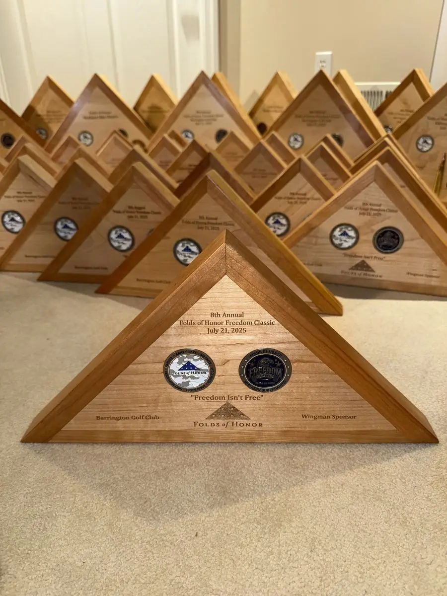 Triangular wooden flag case with two challenge coins and engraved event text, with more cases lined up behind it